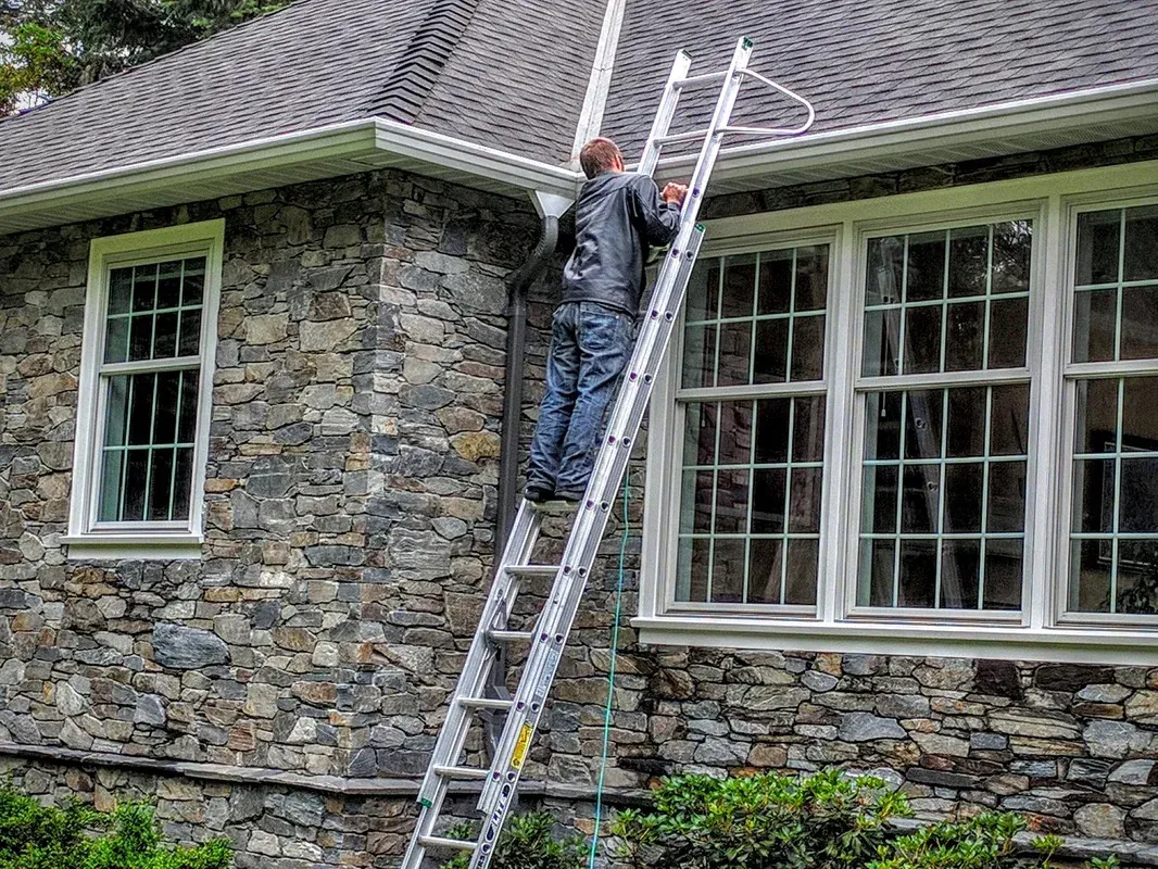 A worker up an extension ladder at the eave of a stone house