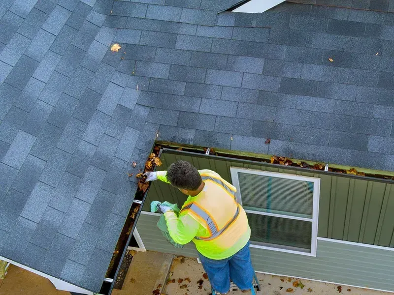 Clearing wet leaves out of a gutter by hand from a ladder set at the eave of a house