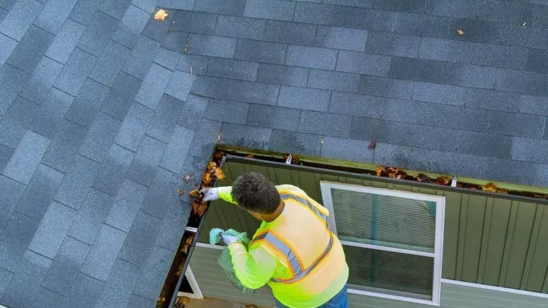 Leaves being scooped from a gutter by a worker in a yellow vest