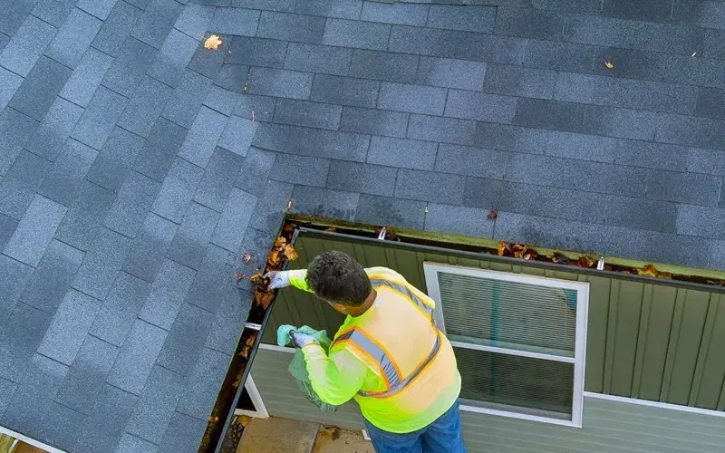A worker in a hi-vis vest scooping wet leaves out of a house gutter